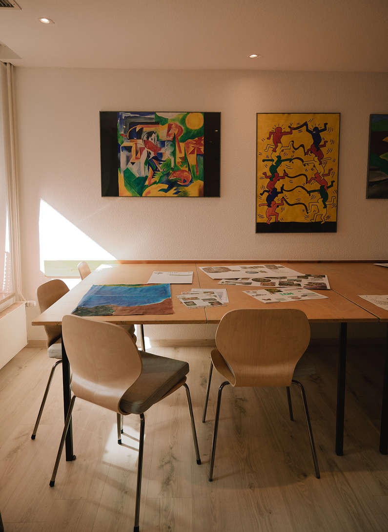 Kreativraum der Klinik Lüneburger Heide mit Tisch, Stühlen und farbenfrohen Bildern an der Wand für Kunsttherapie.